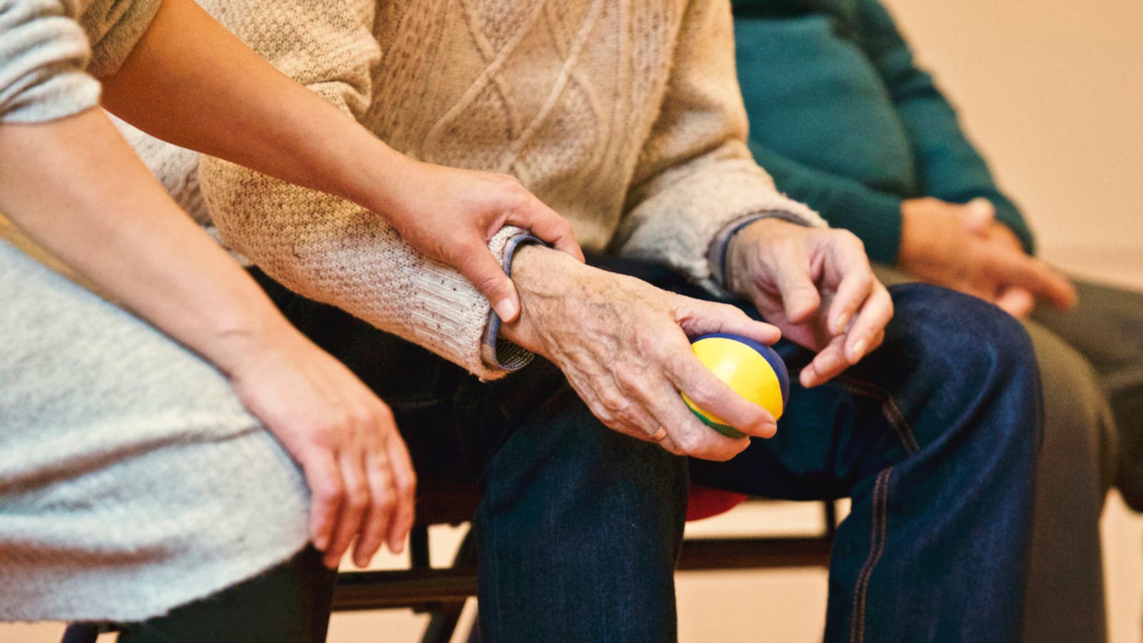 Betreuende Hand stützt behutsam die Hand einer älteren Person mit Therapieball