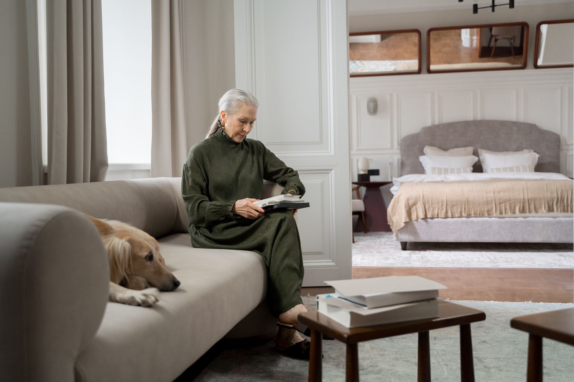 Frau mit grauem Haar liest zu Hause auf dem Sofa, ihr Hund liegt entspannt neben ihr