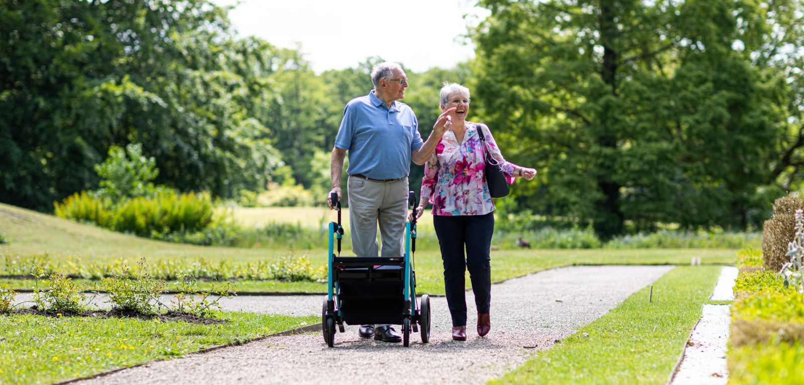 Älteres Paar geht mit einem Rollator gut gelaunt durch einen Park