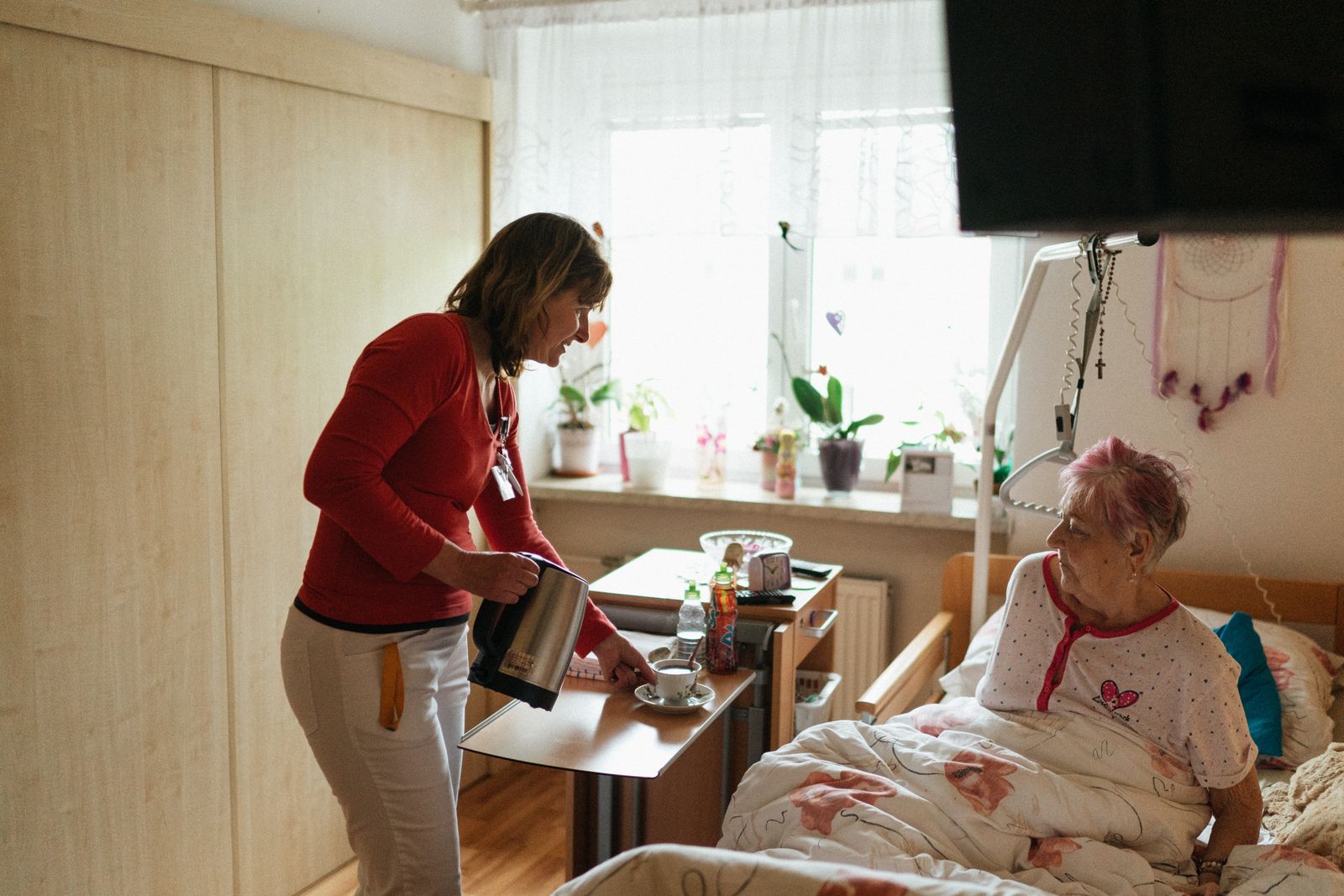 Pflegerin reicht einer älteren Frau im Pflegebett mit Aufrichter eine Tasse Tee – helles, wohnliches Schlafzimmer